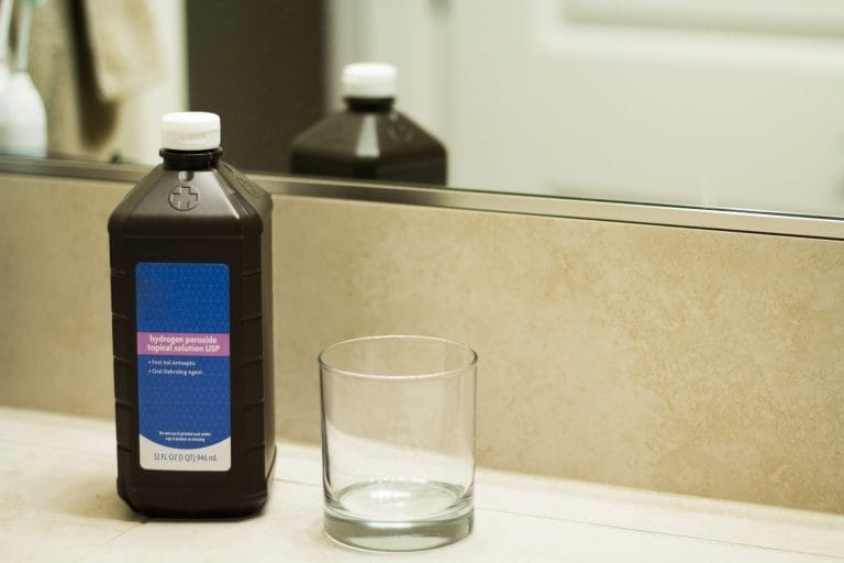 A bottle of hydrogen peroxide next to a glass on a bathroom counter.